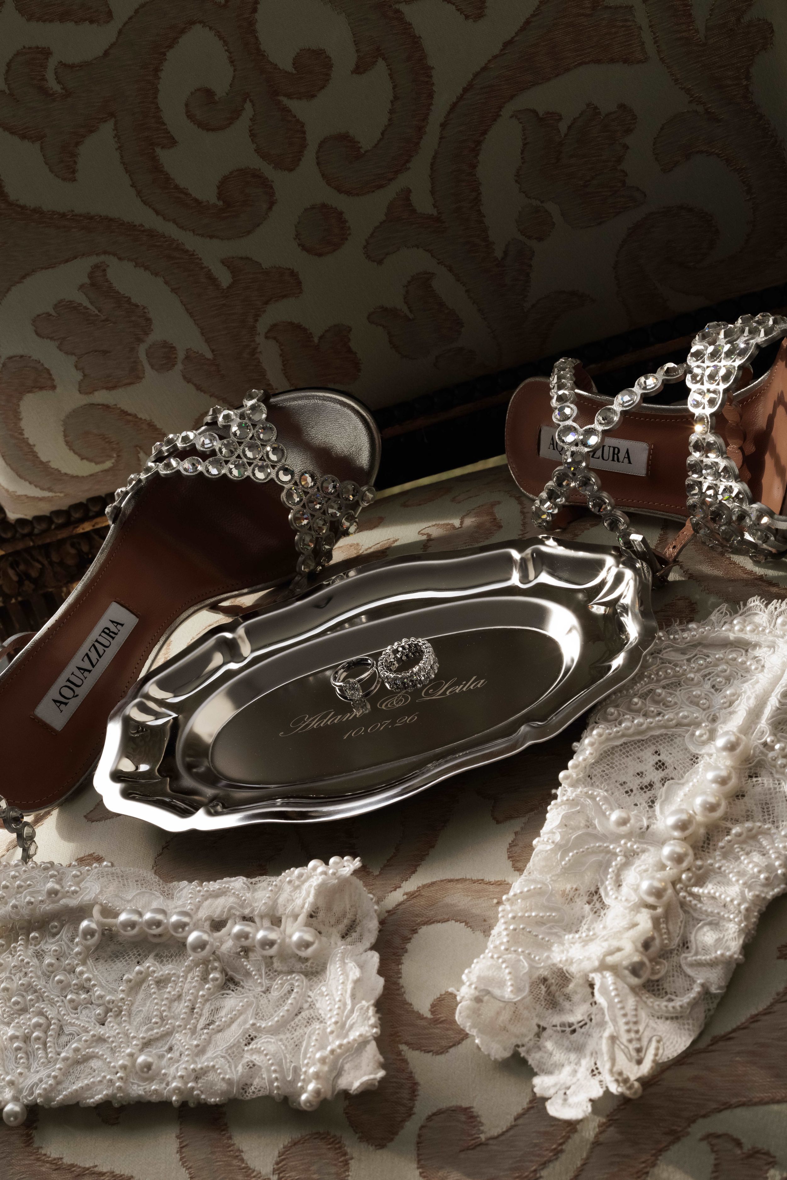 Silver engraved bridal tray with crystal heels, lace garter, and pearl accessories on ornate fabric.