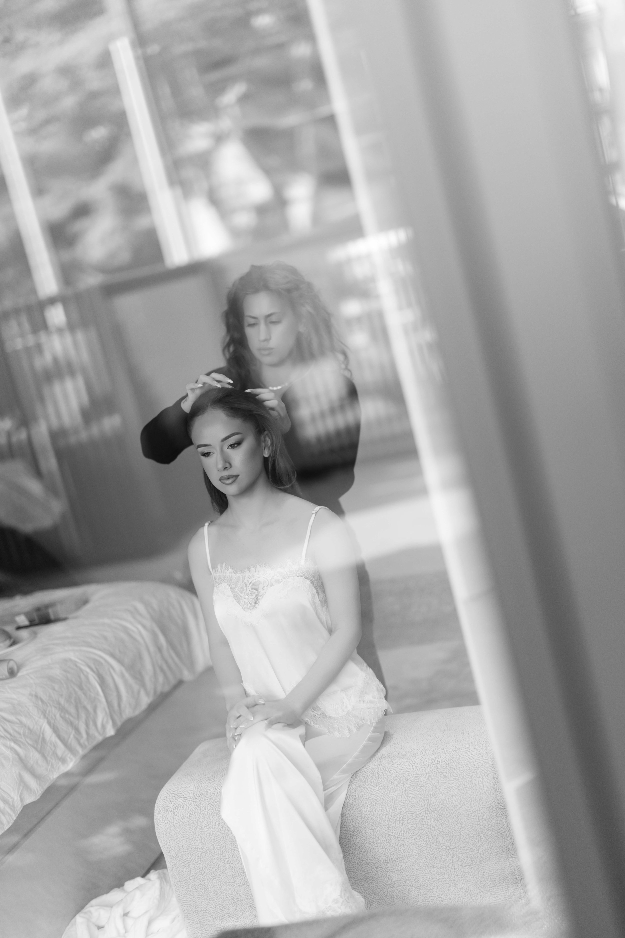 Black and white photo of a bride in white slip dress sitting as hairstylist works on her hair before wedding ceremony.