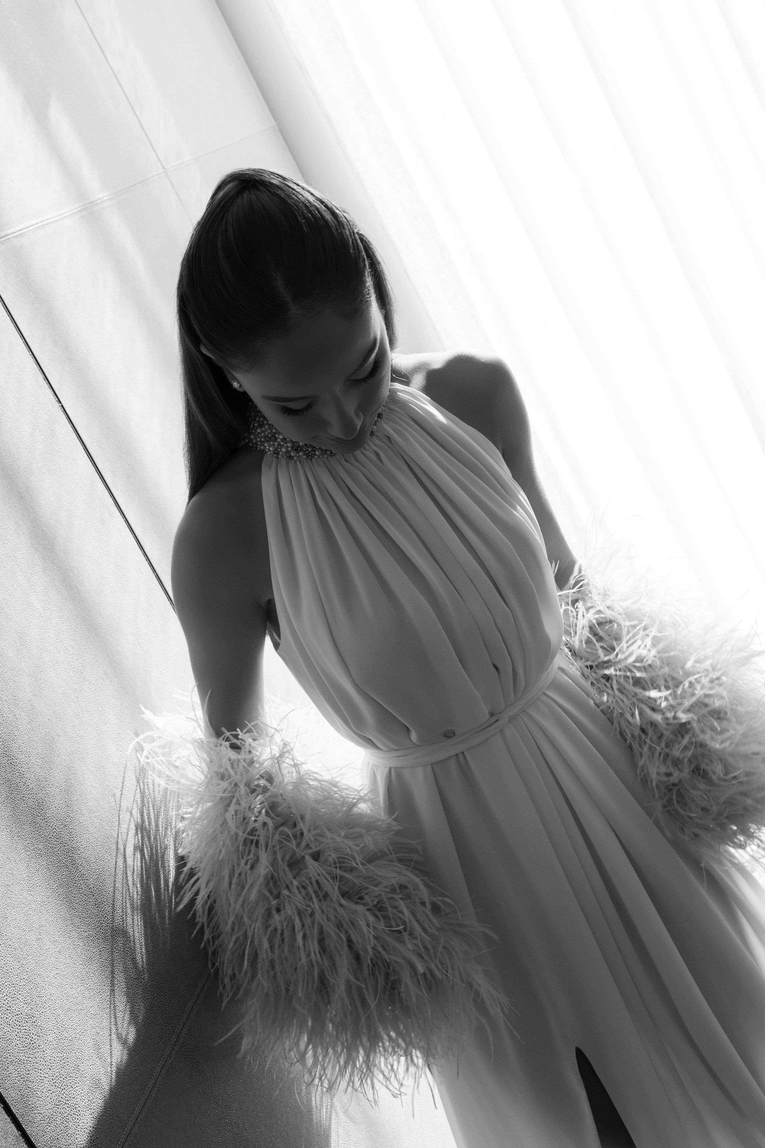 Black and white photo of a woman in an elegant halter gown with feather trim details, posing gracefully indoors.