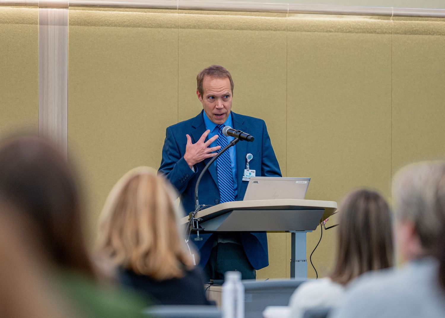A man in a blue suit speaks at a podium with a laptop during a professional conference presentation.