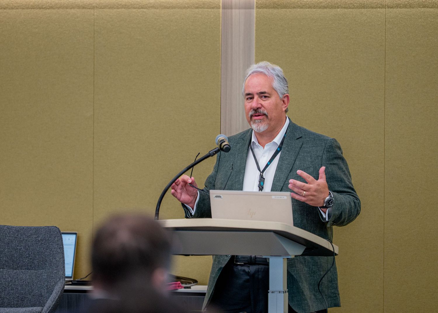 Man in gray blazer speaking at podium with microphone during professional conference presentation.
