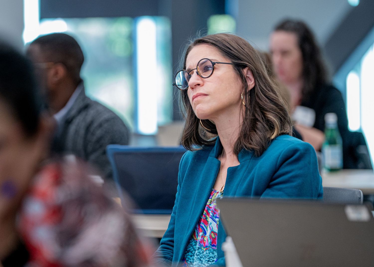 Woman in teal blazer and glasses attentively listening during a professional conference or seminar.