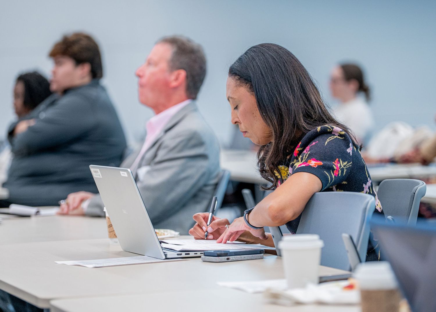 Professional woman taking notes on paper with laptop open during a business conference or corporate training seminar.