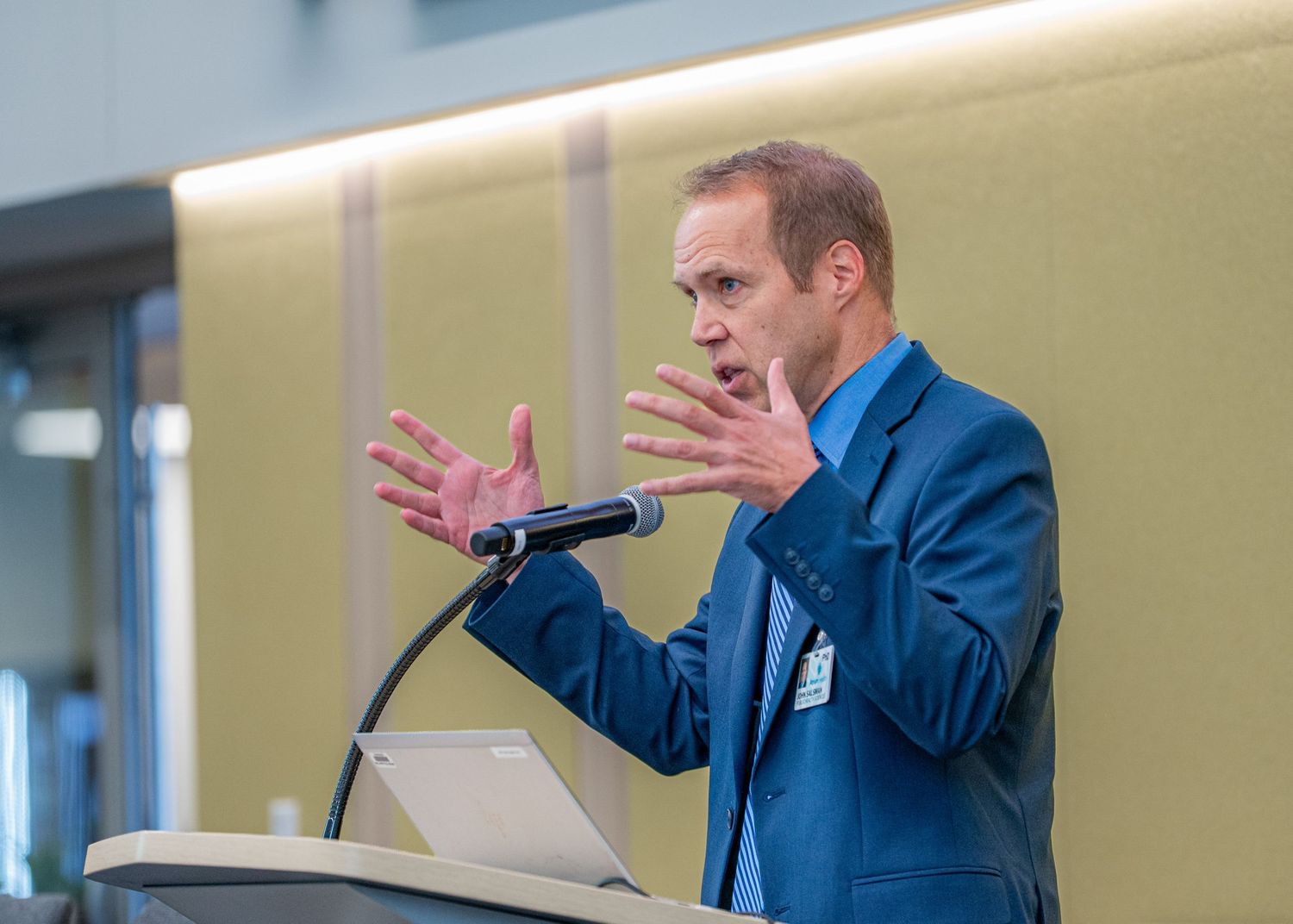 Man in blue suit gestures expressively while speaking at a podium with a microphone during a professional presentation.