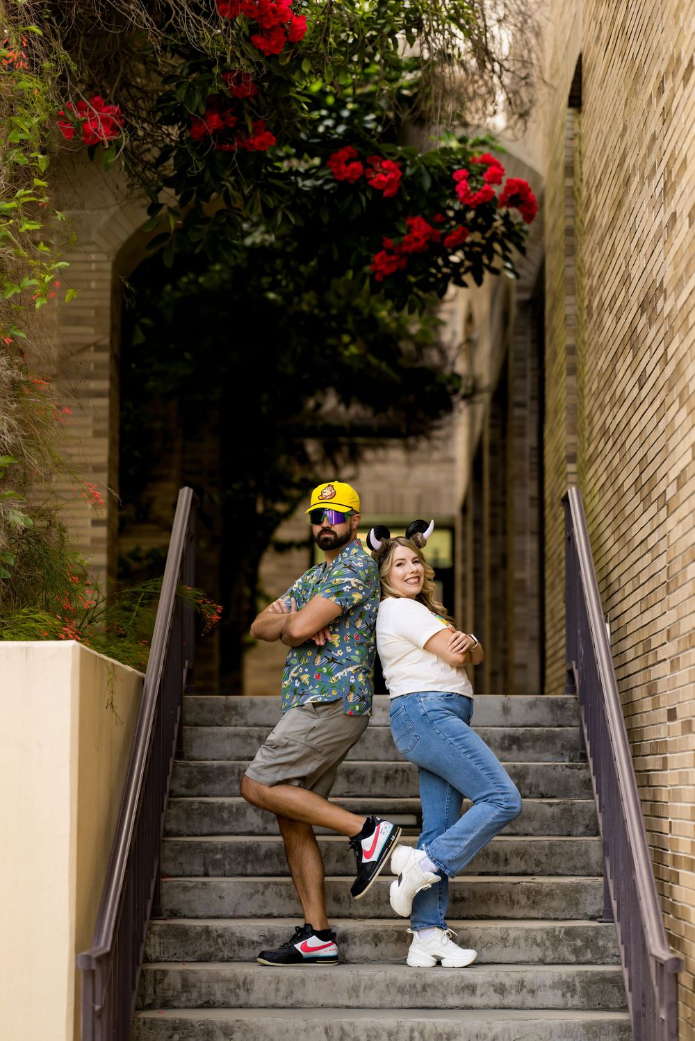 Couple posing back-to-back on outdoor stairs, man in yellow cap and floral shirt, woman in jeans, surrounded by red flowers.