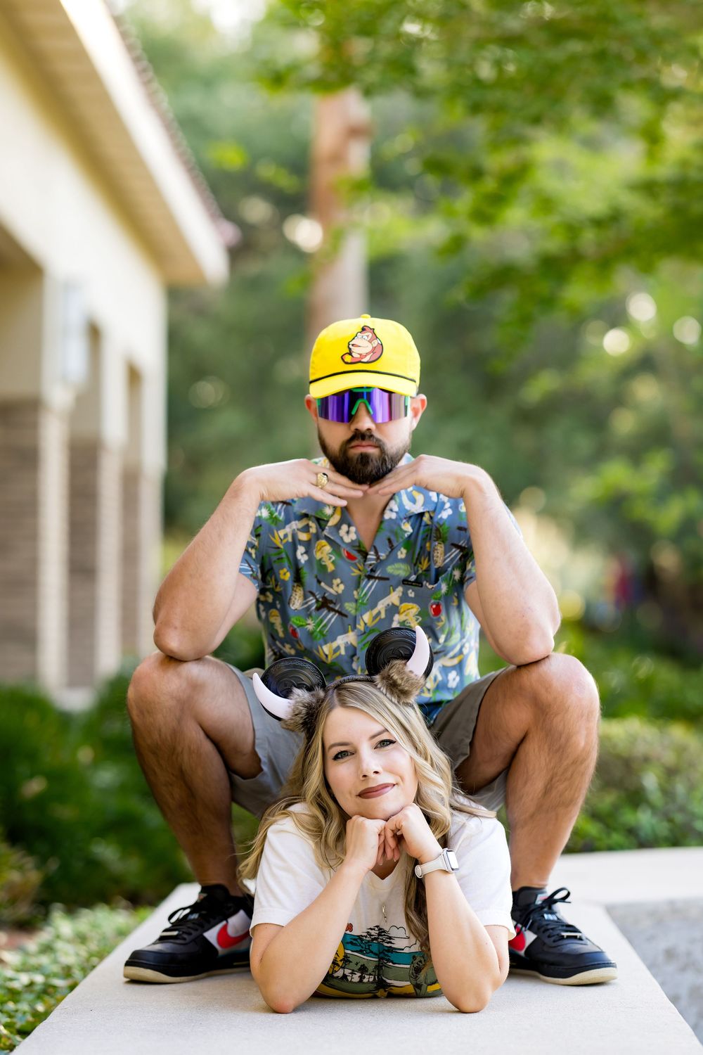 A bearded man in a yellow cap and sunglasses squats behind a smiling blonde woman lying on a pathway outdoors.