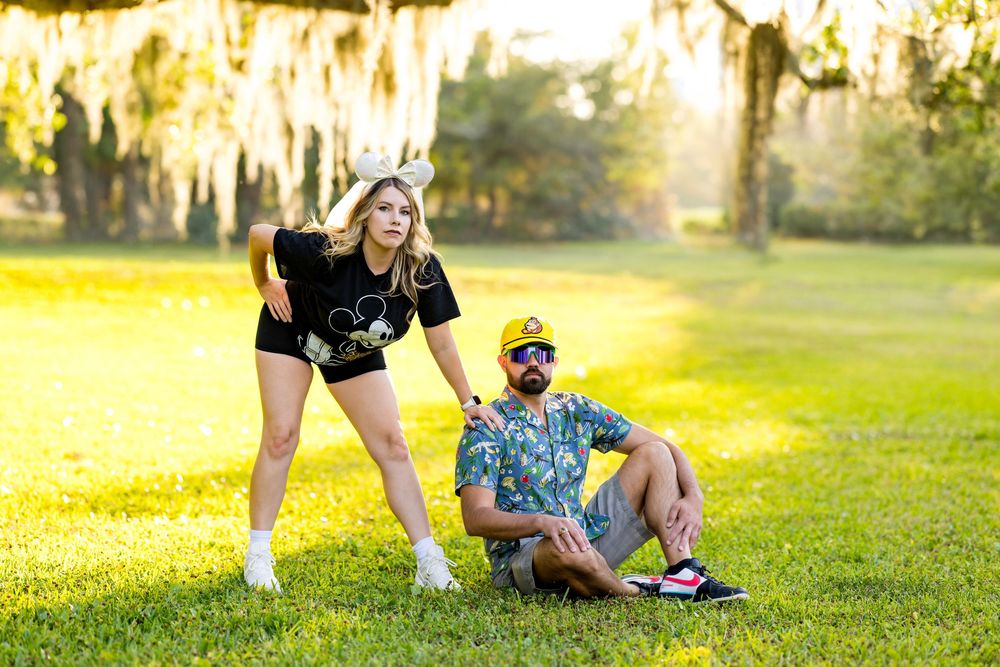 Couple posing outdoors on green grass under Spanish moss-draped trees, woman in black Disney shirt, man in floral shirt.