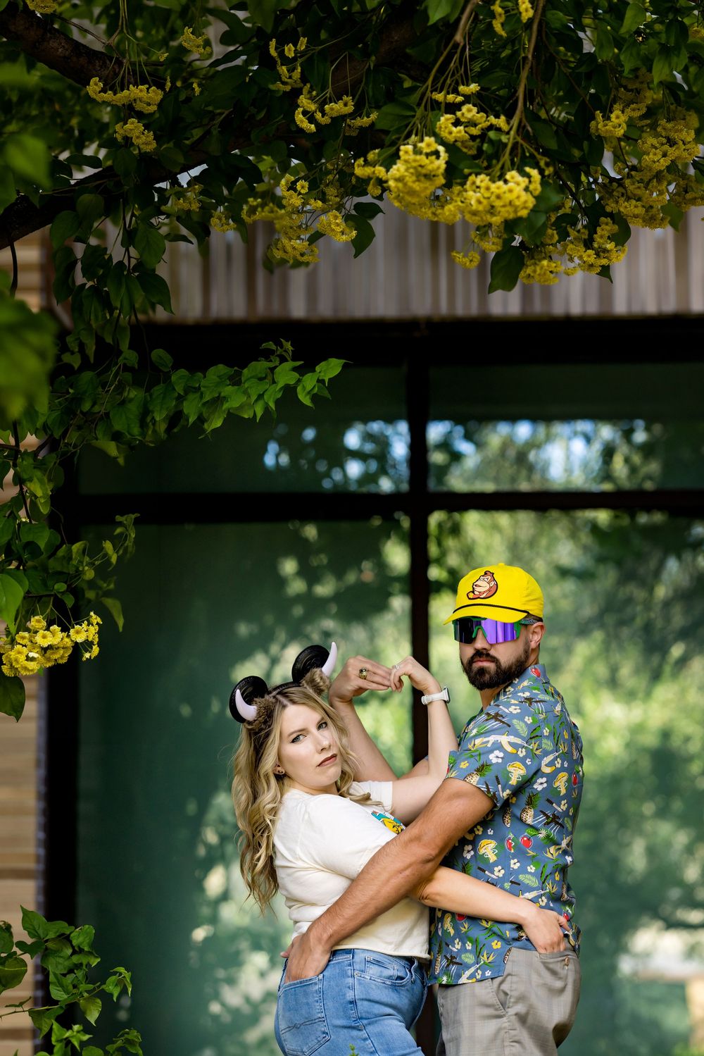 Couple posing under yellow flowering tree, woman with mouse ears and man in yellow cap and floral shirt.