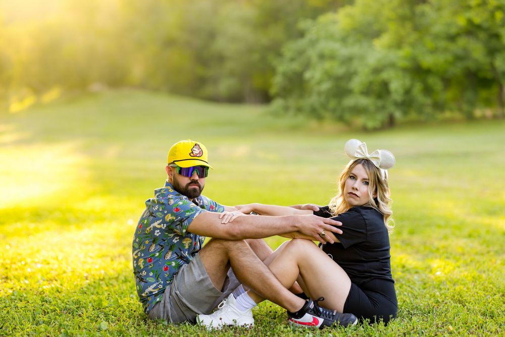 Couple sitting on green grass in sunny park, man in yellow cap and floral shirt, woman in black outfit with bow headband.