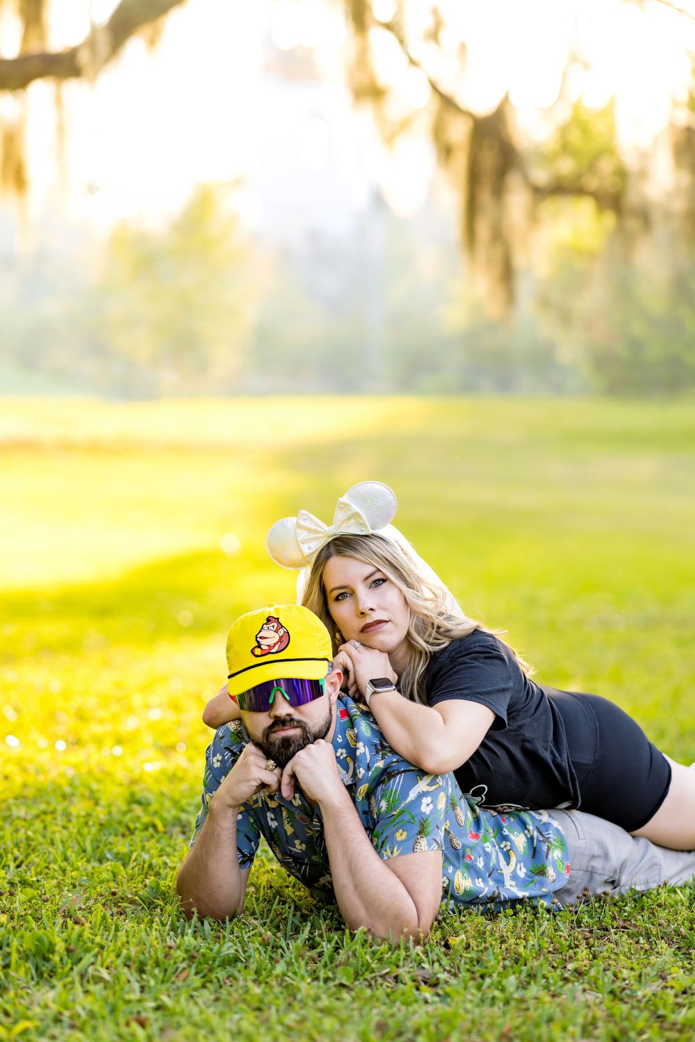 Couple posing on green grass under sunlit trees, man in yellow cap and sunglasses, woman in black top with mouse ears.