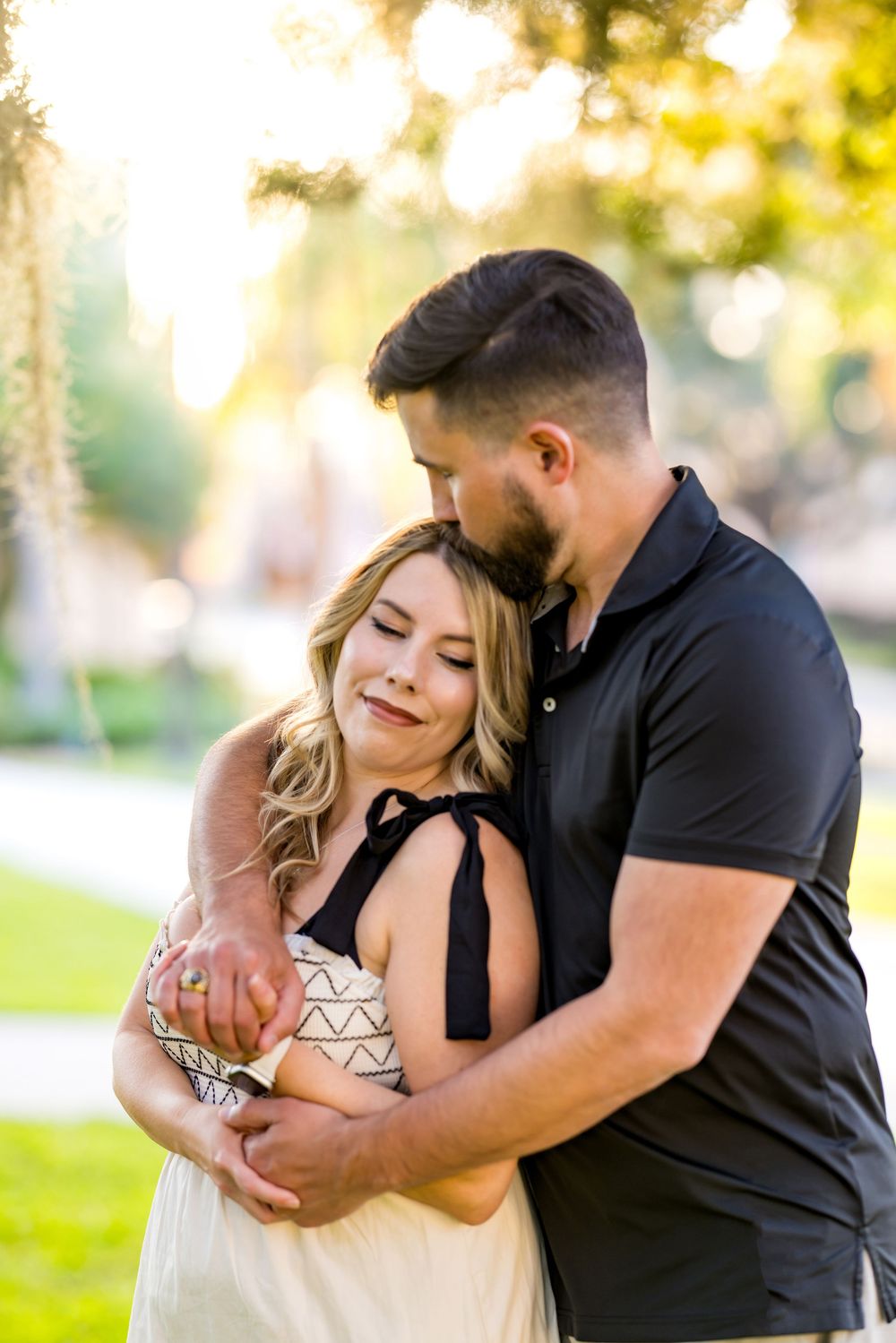 A couple embraces outdoors, man in black shirt kisses womans head as she smiles, golden sunlight filters through trees.