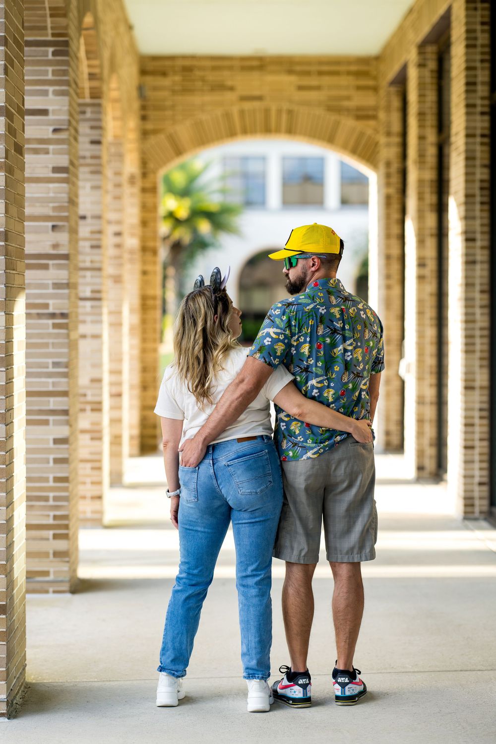 Couple embracing from behind in a brick archway corridor, woman in jeans and man in colorful shirt and yellow cap.