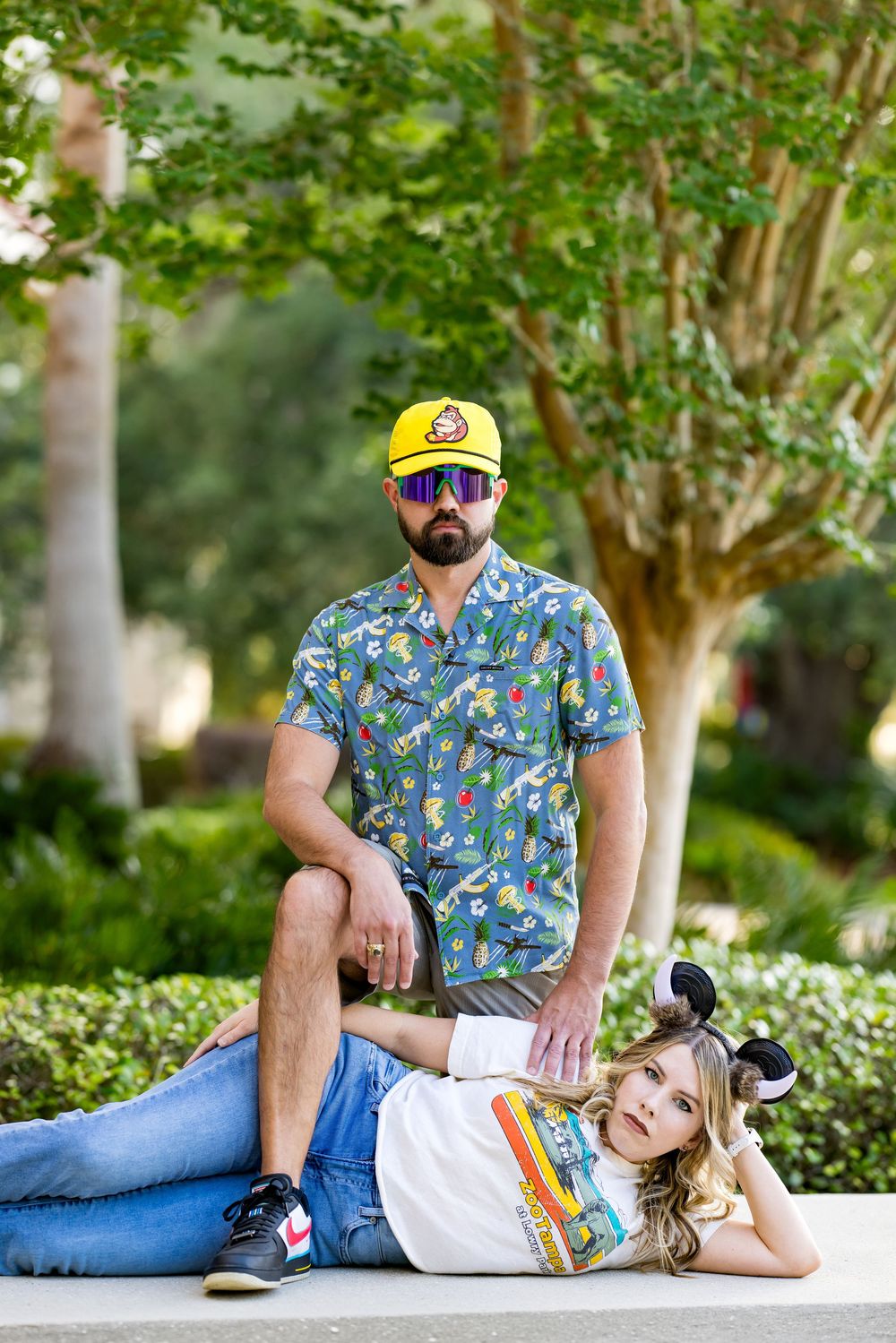 Couple posing outdoors, man in yellow cap and Hawaiian shirt standing over woman in Mickey ears lying on ground.