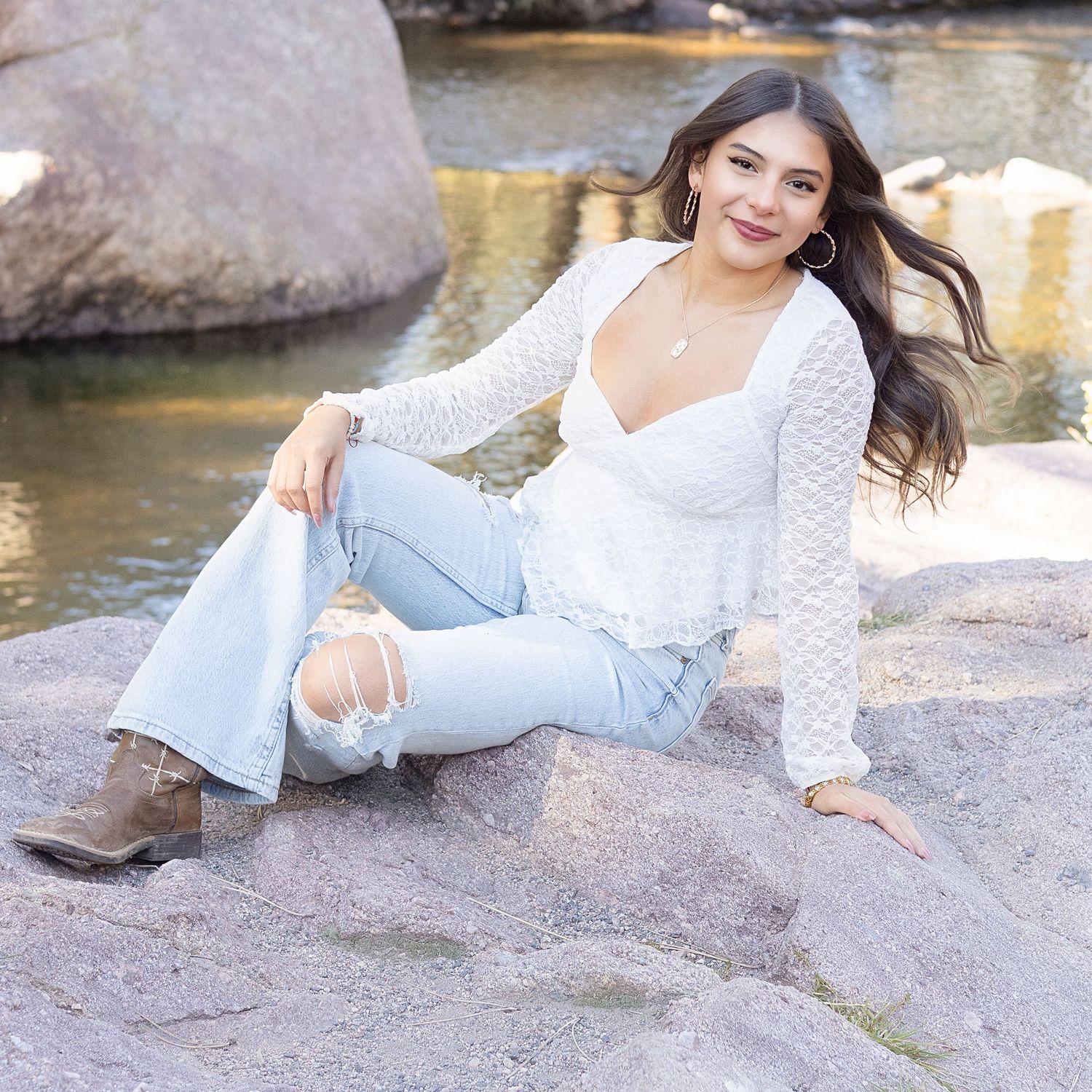 Young woman in white lace top and ripped jeans sitting on rocks near a river, wearing brown cowboy boots.