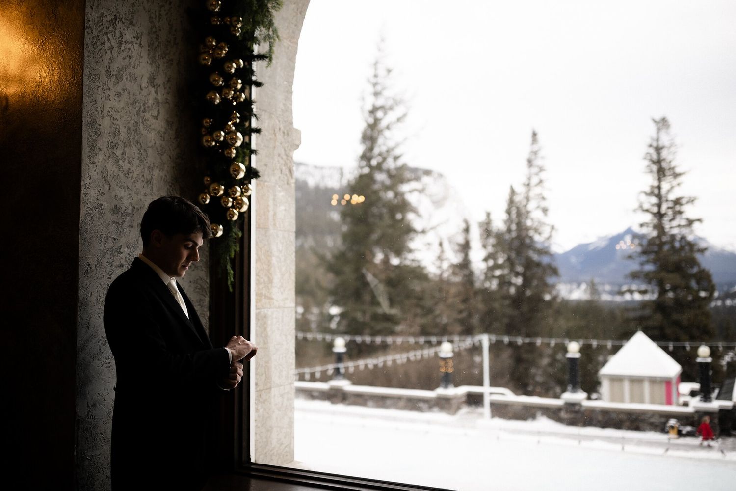Groom fastening cufflinks at a Fairmont Banff Springs window with gold ornament garland and snowy mountains outside