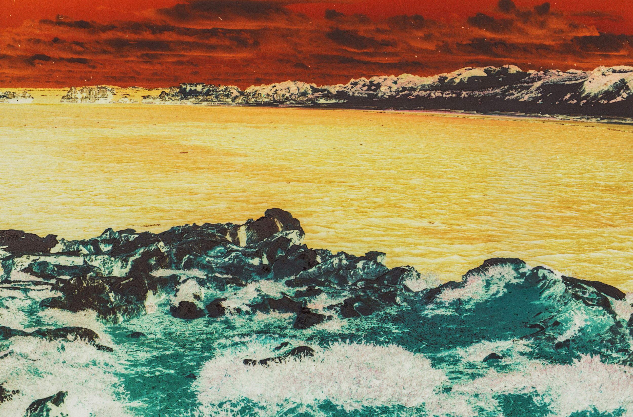 Surreal infrared seascape with teal ocean waves crashing on rocks under a vivid orange and red sky.