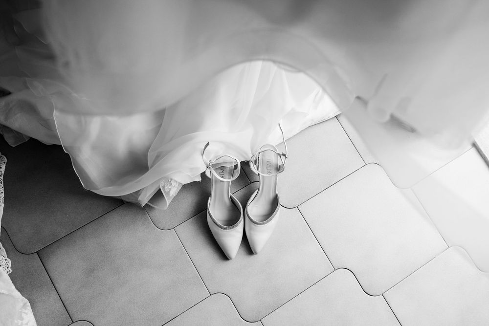 Black and white photo of elegant bridal heels and glasses placed on tiled floor beneath a flowing white wedding dress.