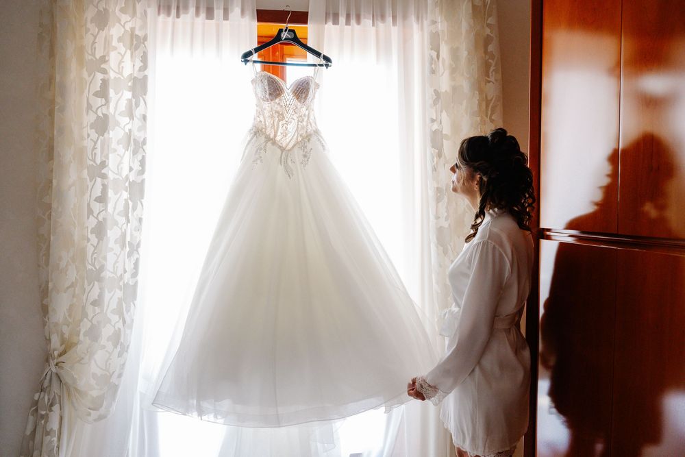 Bride in white robe admiring elegant ball gown wedding dress hanging by sunlit window before ceremony.