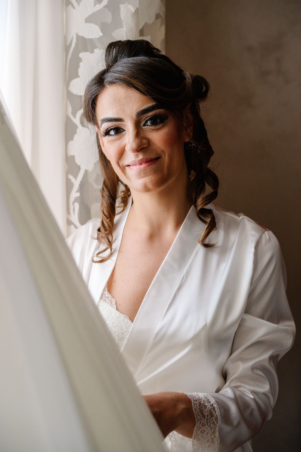 Smiling bride in white satin robe holding wedding dress near window with floral curtains, elegant updo hairstyle.
