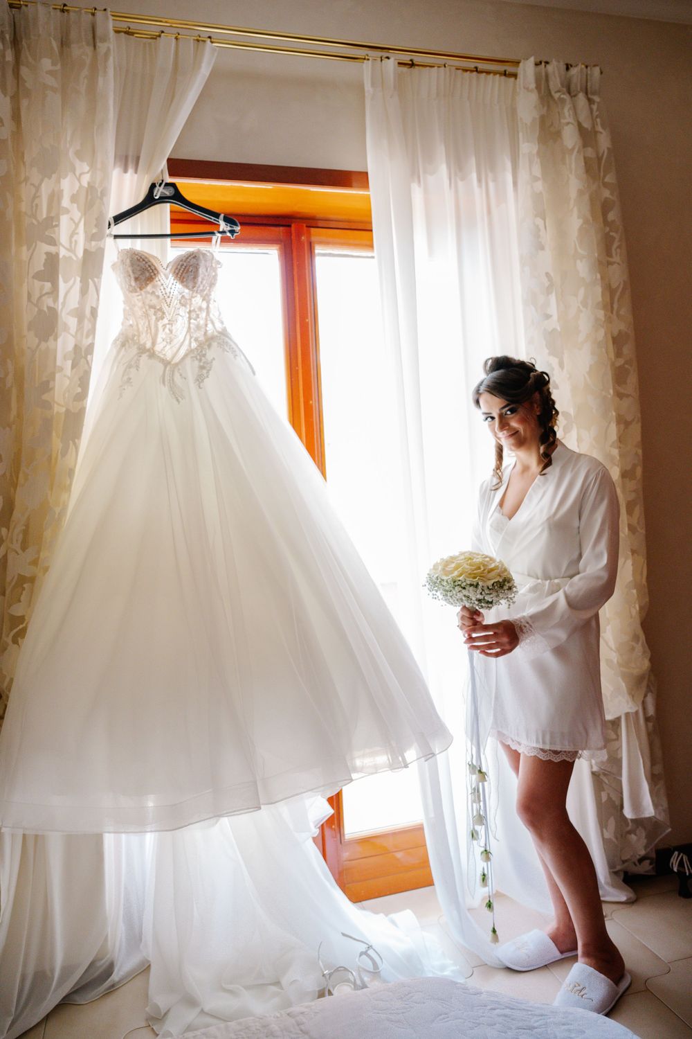 Bride in white robe admires her elegant lace ball gown wedding dress hanging by a sunlit window with sheer curtains.