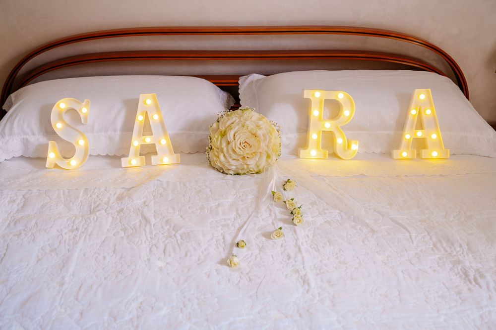 Illuminated marquee letters spelling SARA on a white bed with a yellow rose bouquet, perfect for a bridal room decoration.