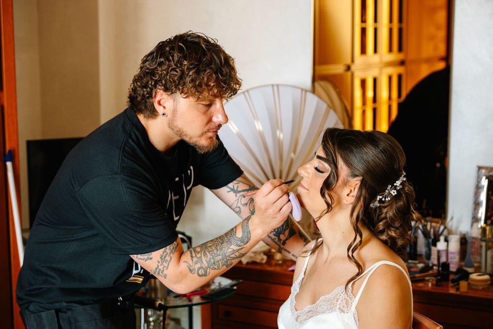 Tattoed makeup artist applying makeup to a bride wearing a white dress and floral hair accessory.
