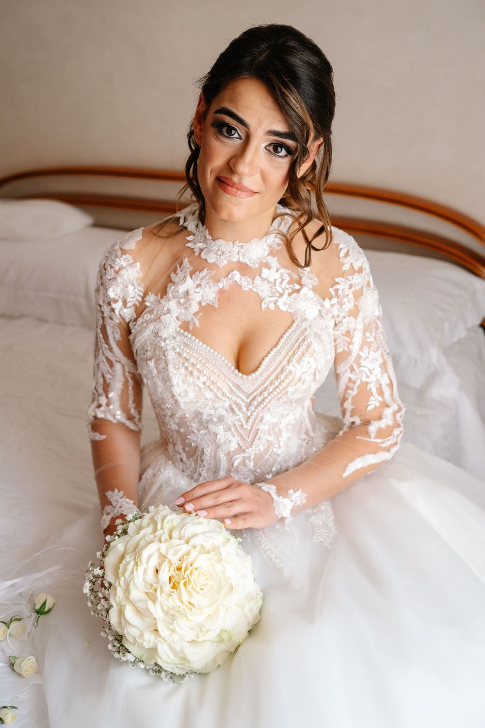 Bride in lace wedding gown holding white peony bouquet, seated on bed with elegant updo and dramatic makeup.
