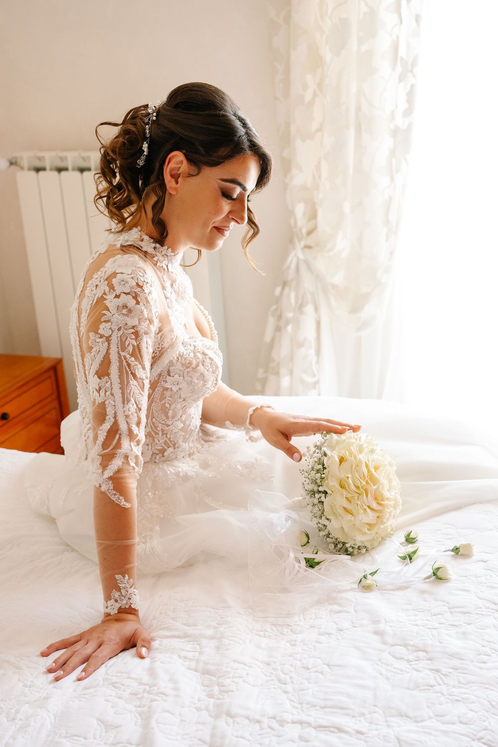 Elegant bride in lace wedding dress arranging white flower bouquet on bed near window.