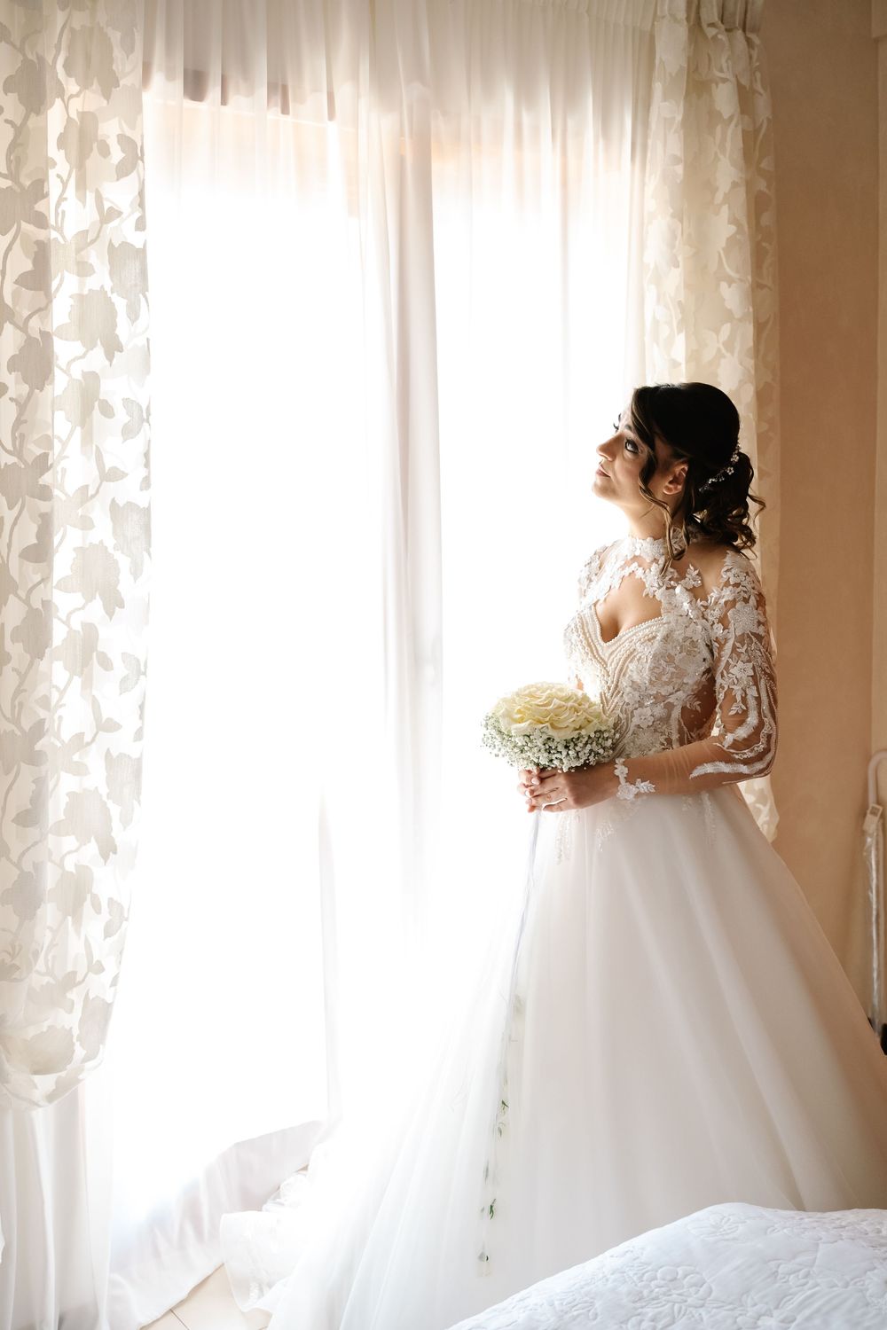 Bride in lace wedding gown holding bouquet standing by bright window, smiling softly in elegant bridal portrait.