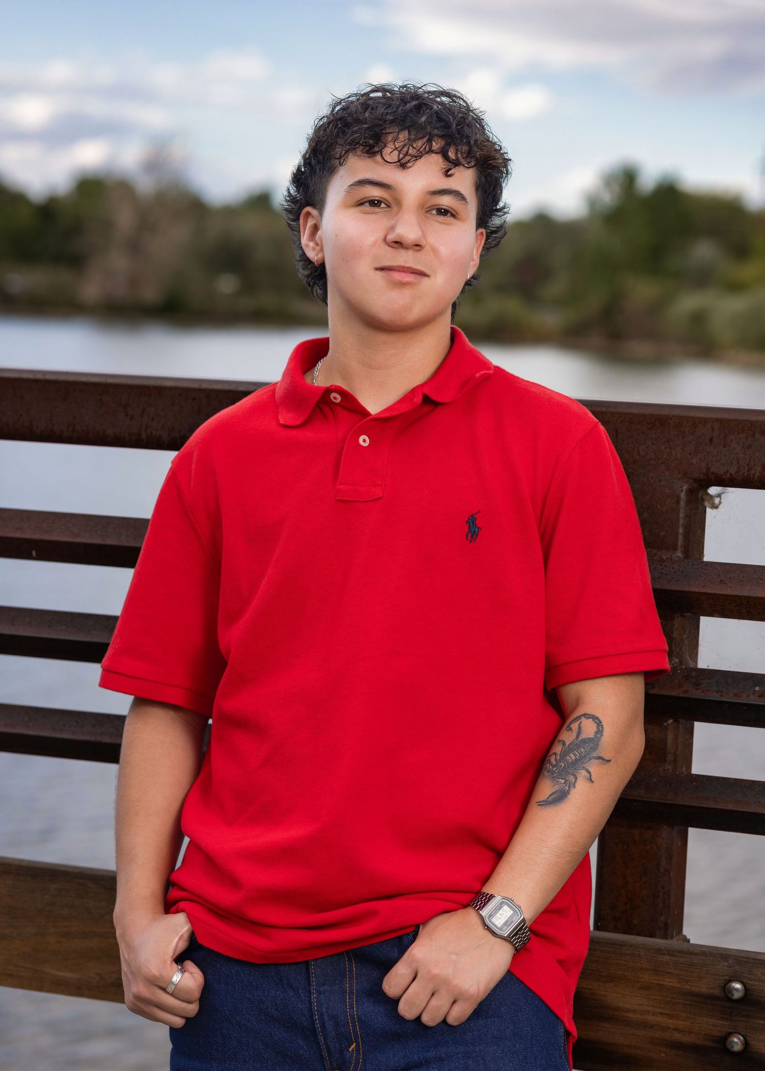 Young man in red Polo shirt with tattoo poses on wooden bridge near water for portrait photo.
