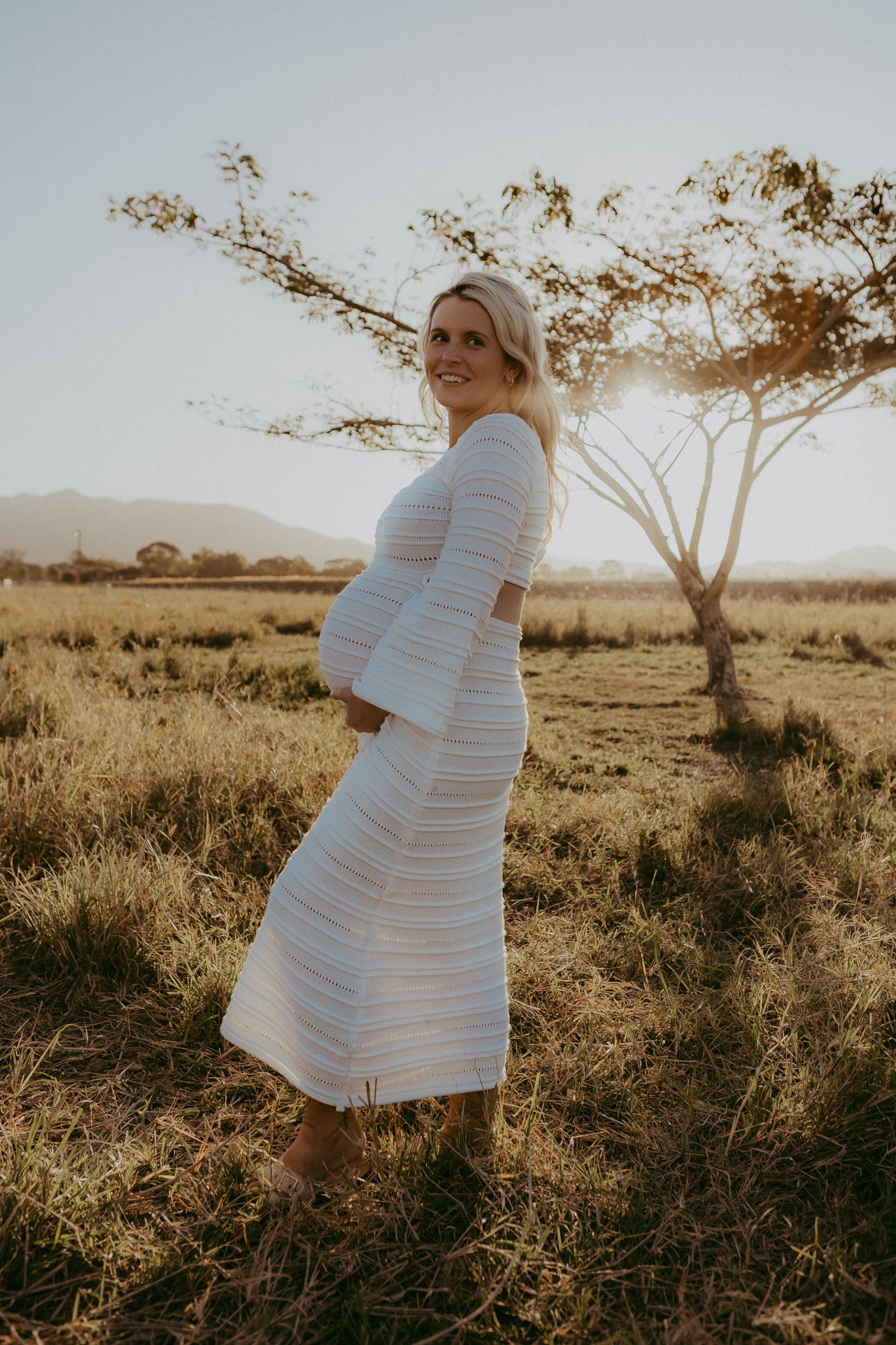 A pregnant woman in a white striped dress stands in a golden sunlit field with a tree behind her.