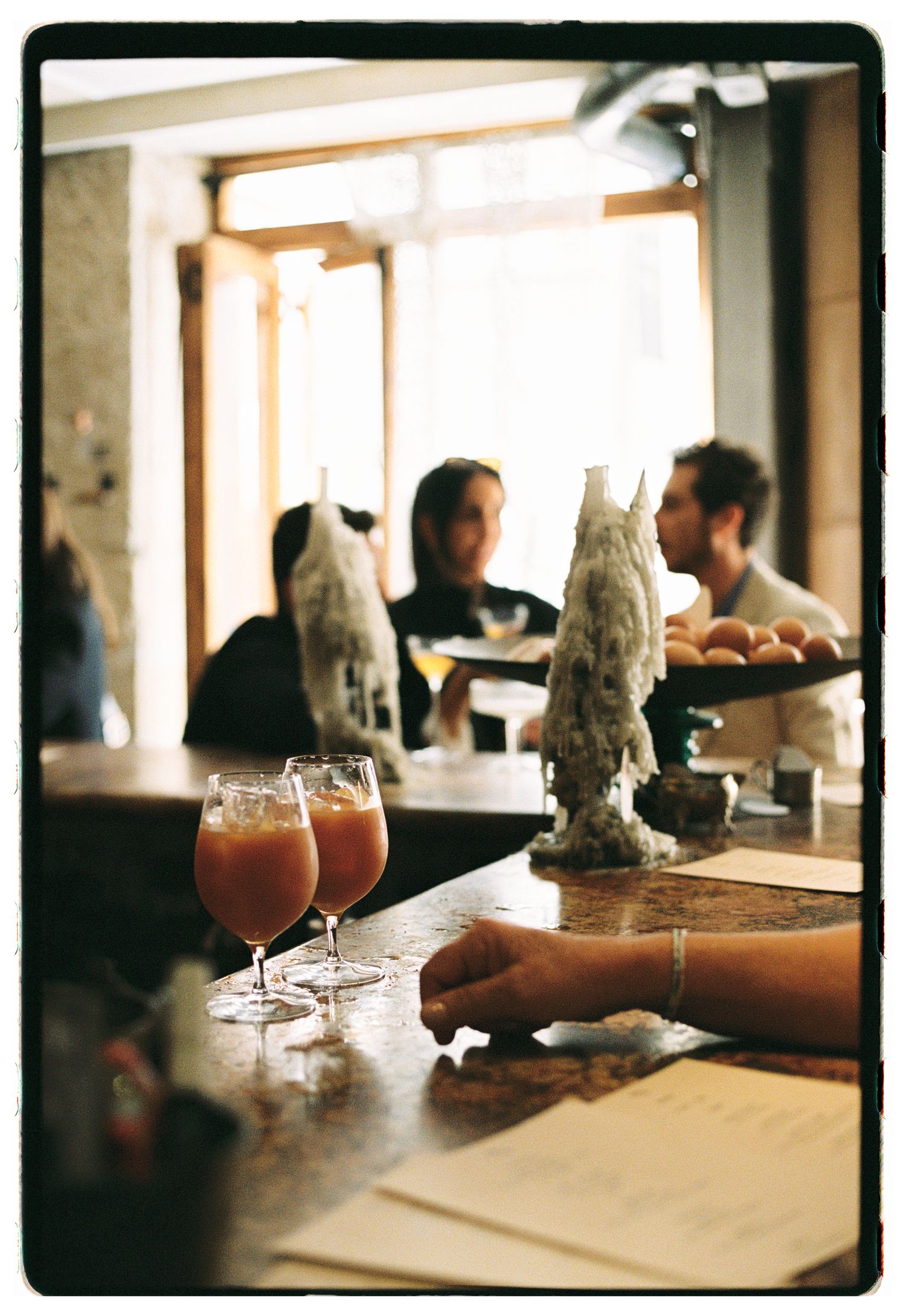 Two red cocktails on a marble bar with blurred patrons and warm backlight in a cozy restaurant setting. paris film photographer