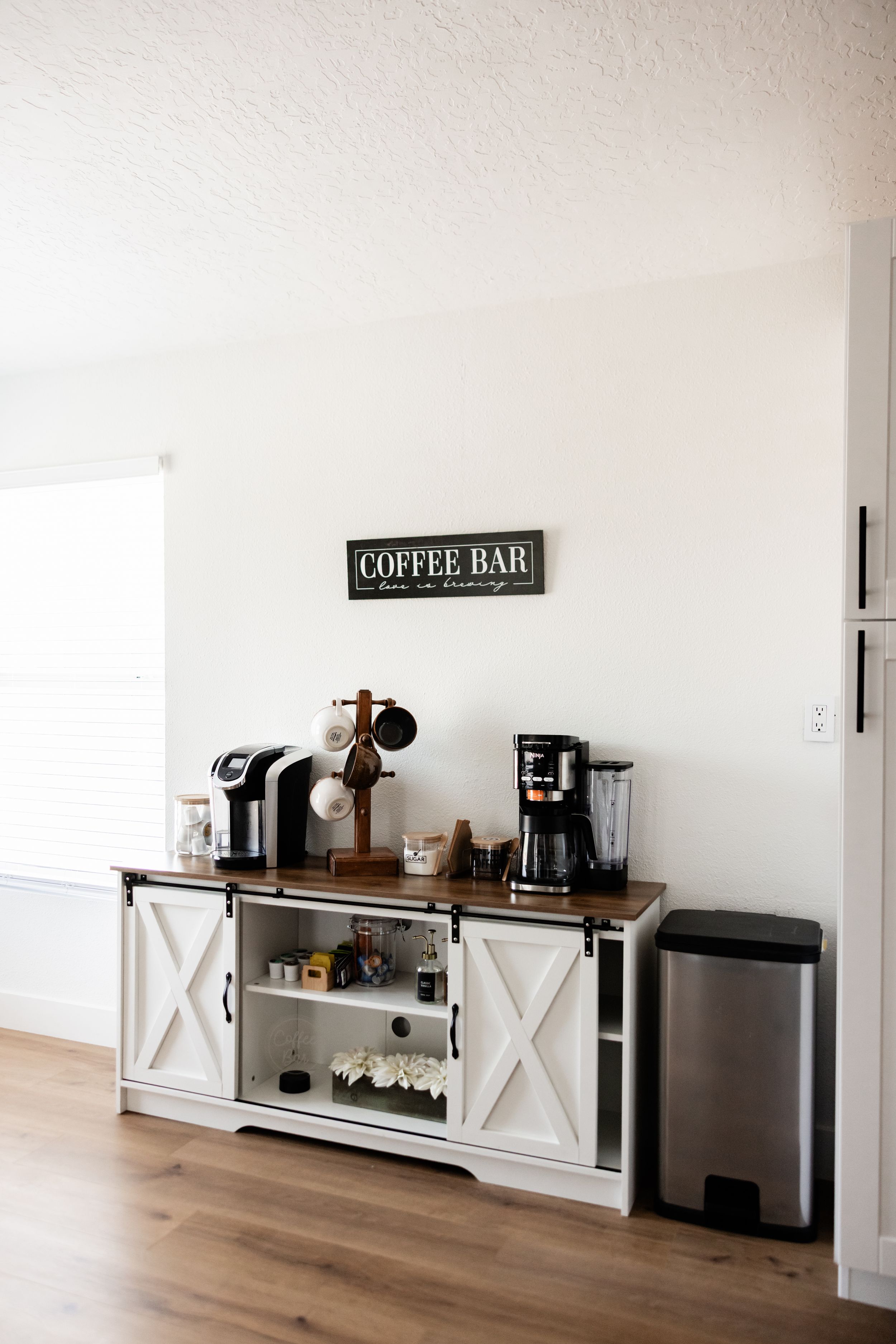 Farmhouse-style white coffee bar cabinet with Keurig, drip coffee maker, blender, and decorative Coffee Bar sign.