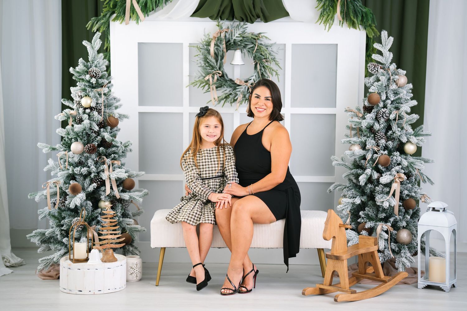Woman and young girl pose together on bench surrounded by festive Christmas trees in decorated holiday studio.