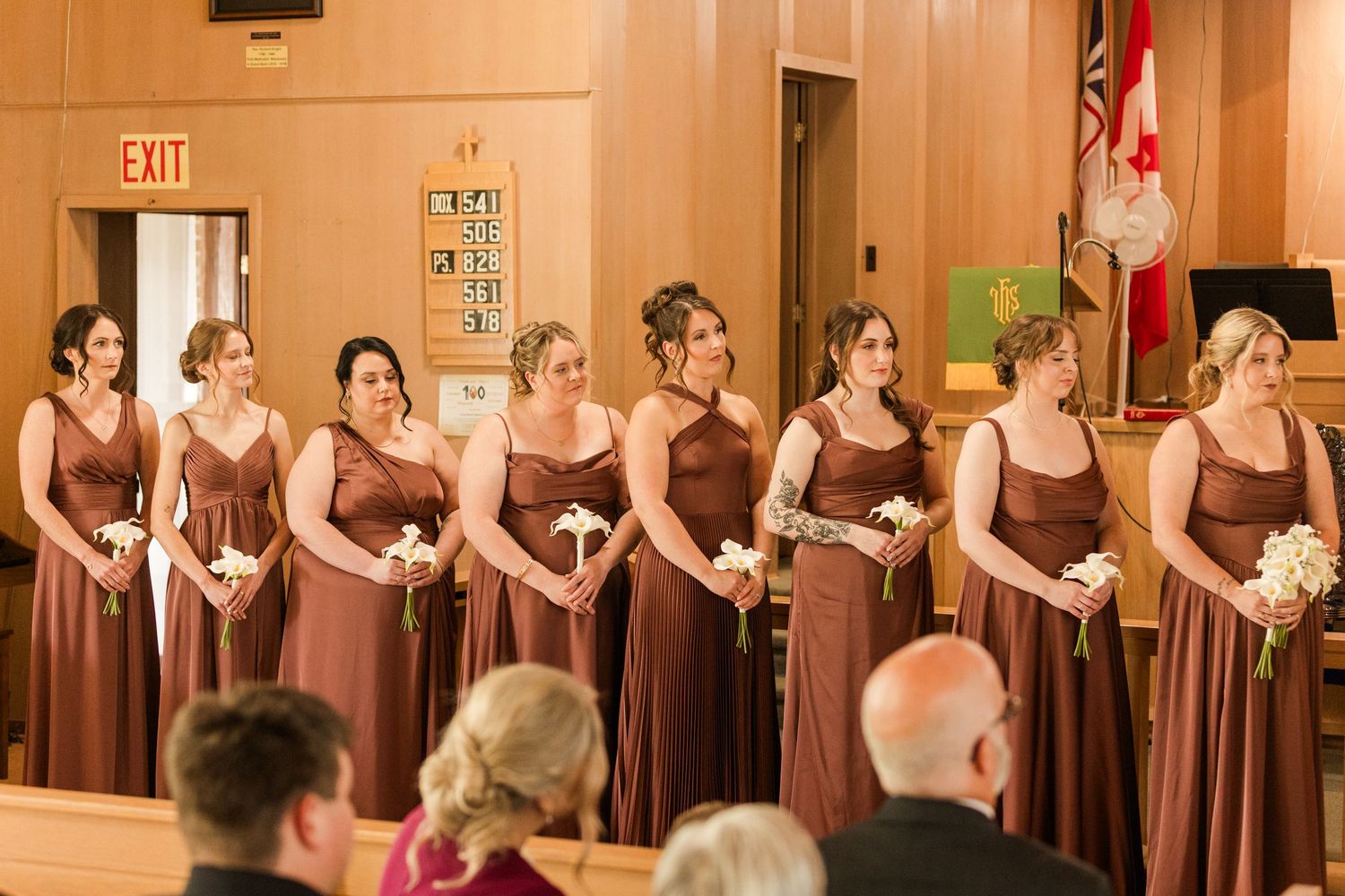 Eight bridesmaids in matching brown dresses holding white bouquets stand in a row during a wedding ceremony.