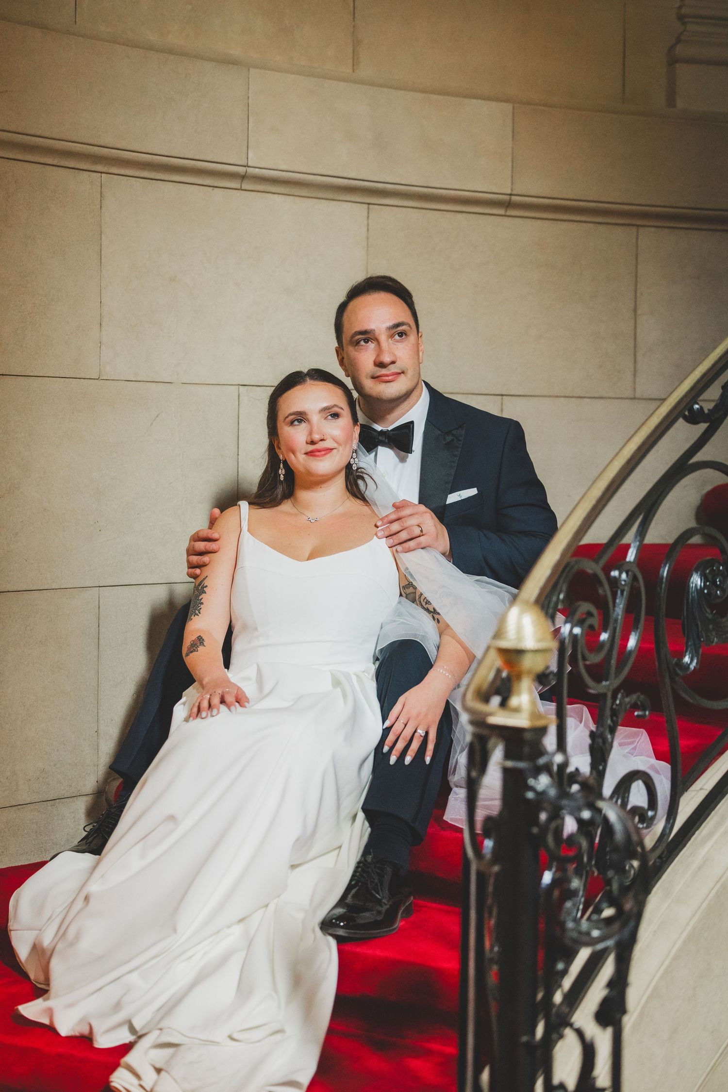 Bride and groom sitting on red-carpeted staircase, she in white gown, he in dark tuxedo with bow tie. Flash wedding photography at meridian