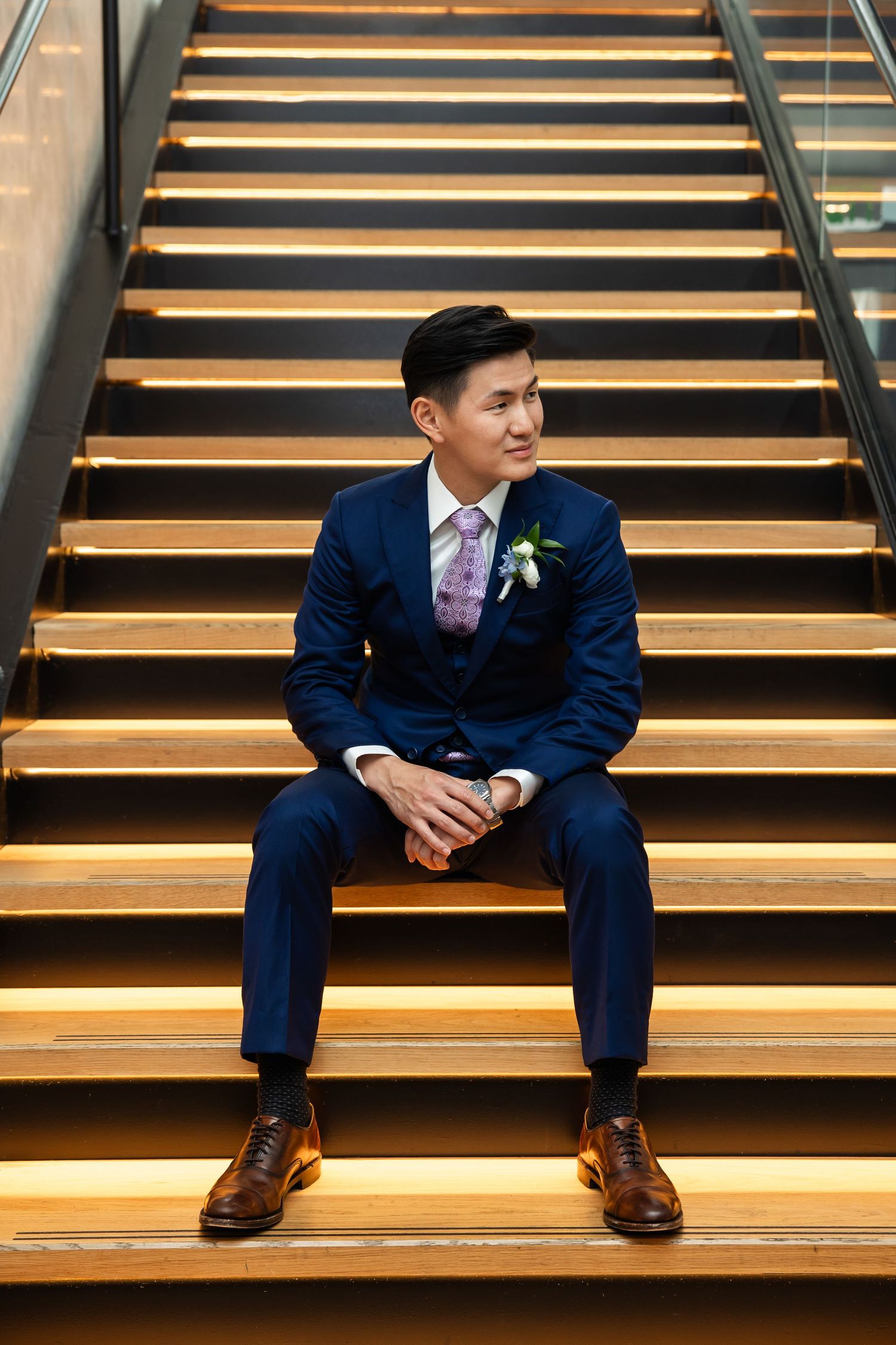 A groom in a navy suit with boutonniere sits on illuminated wooden stairs, looking away thoughtfully.