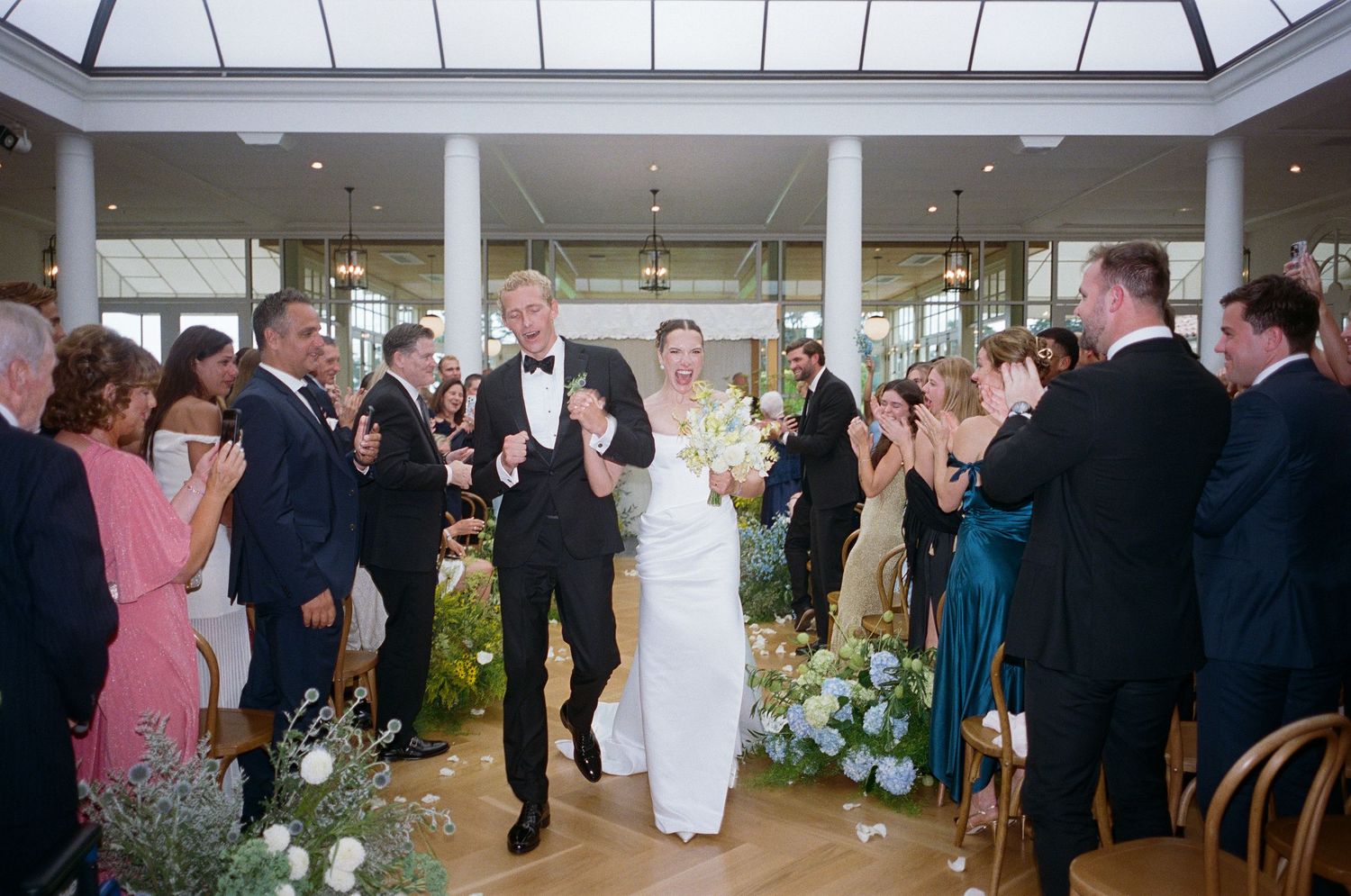 Bride and groom walk down the aisle as guests celebrate at an elegant indoor wedding ceremony.