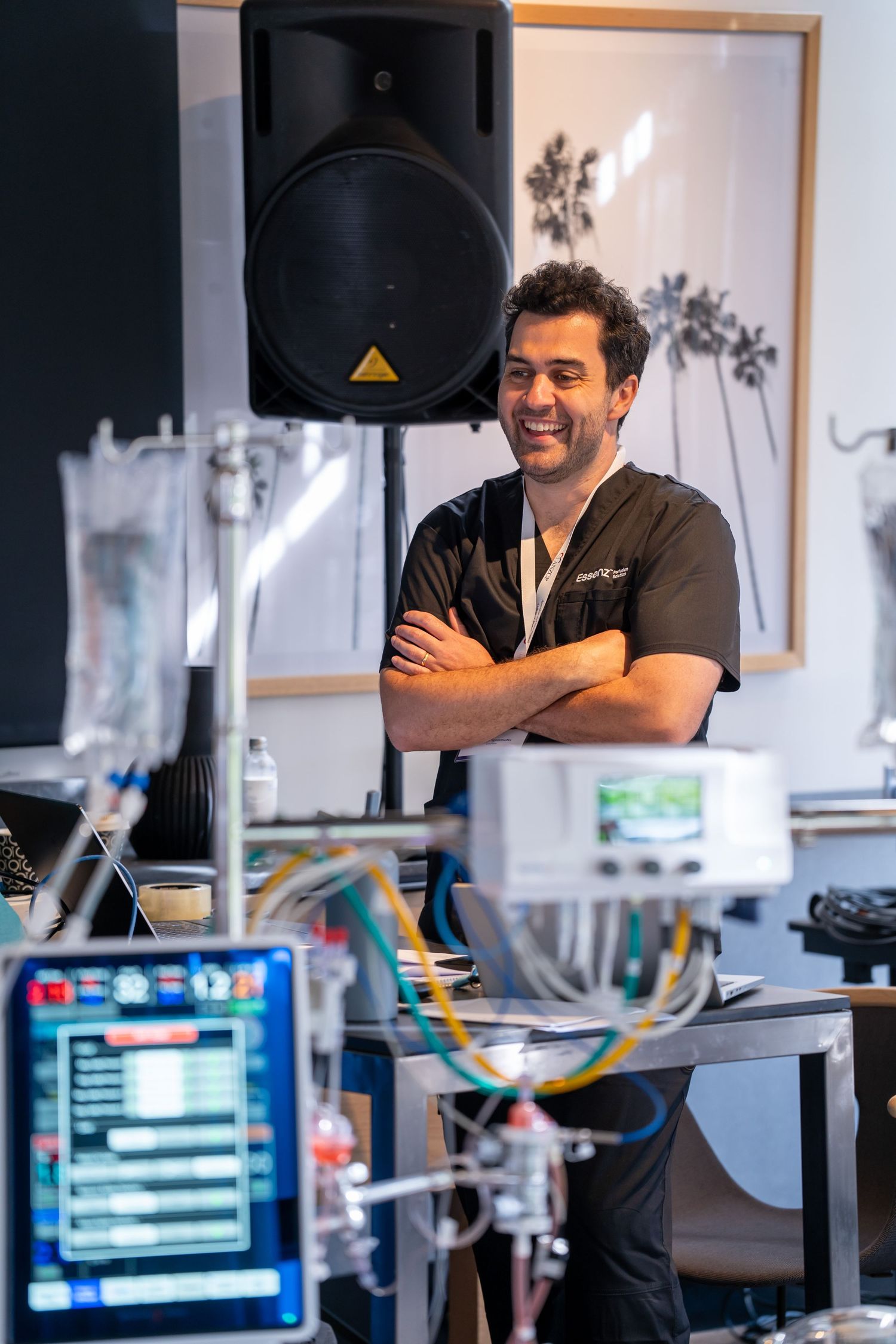 Smiling male medical professional in scrubs stands with arms crossed near medical equipment and monitors.