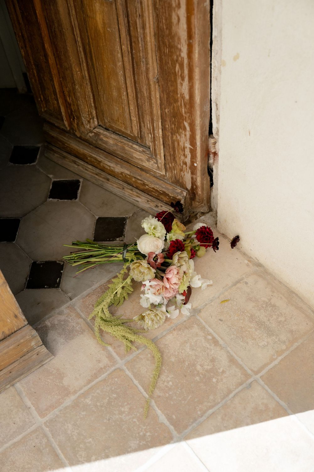 Bridal bouquet with pink and burgundy flowers resting on tiled floor beside rustic wooden door.