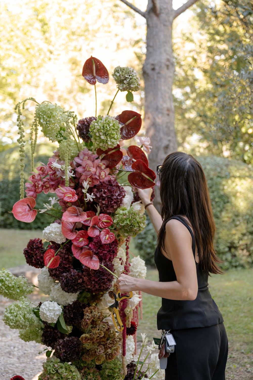Woman in black dress arranging a large floral display with red anthuriums and hydrangeas outdoors.