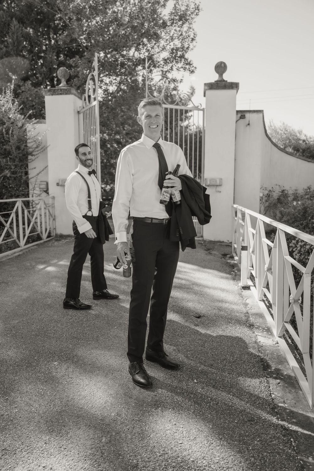 Two groomsmen in formal attire walk through elegant white gates on a sunny wedding day.