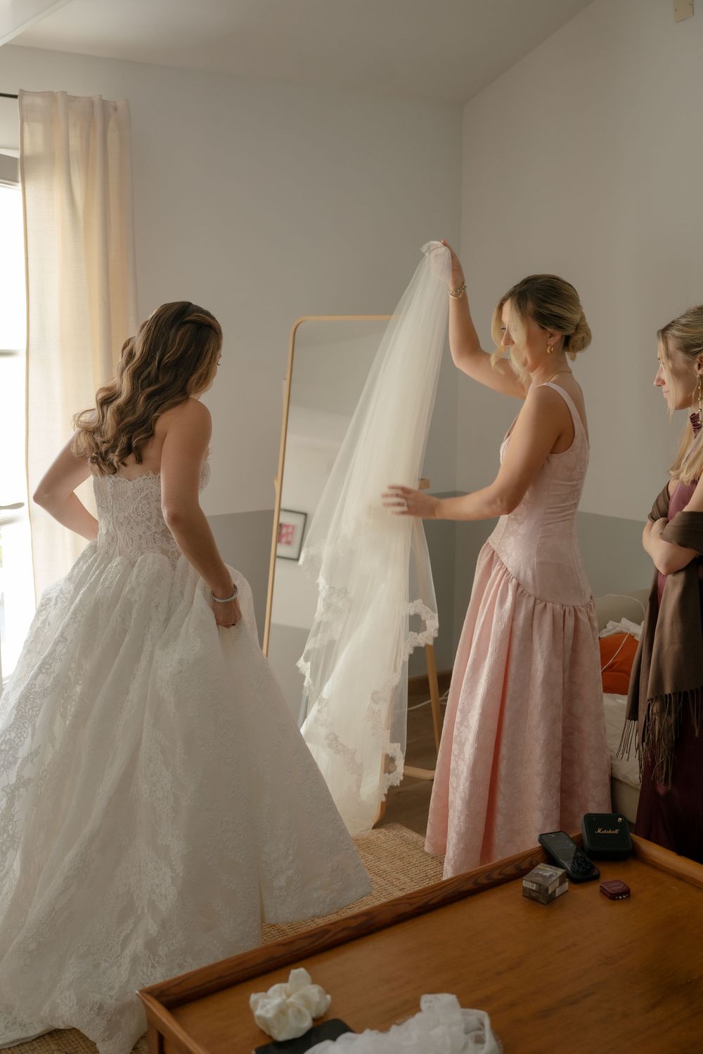 Bridesmaid in pink dress helps bride with white lace wedding gown and veil before ceremony.