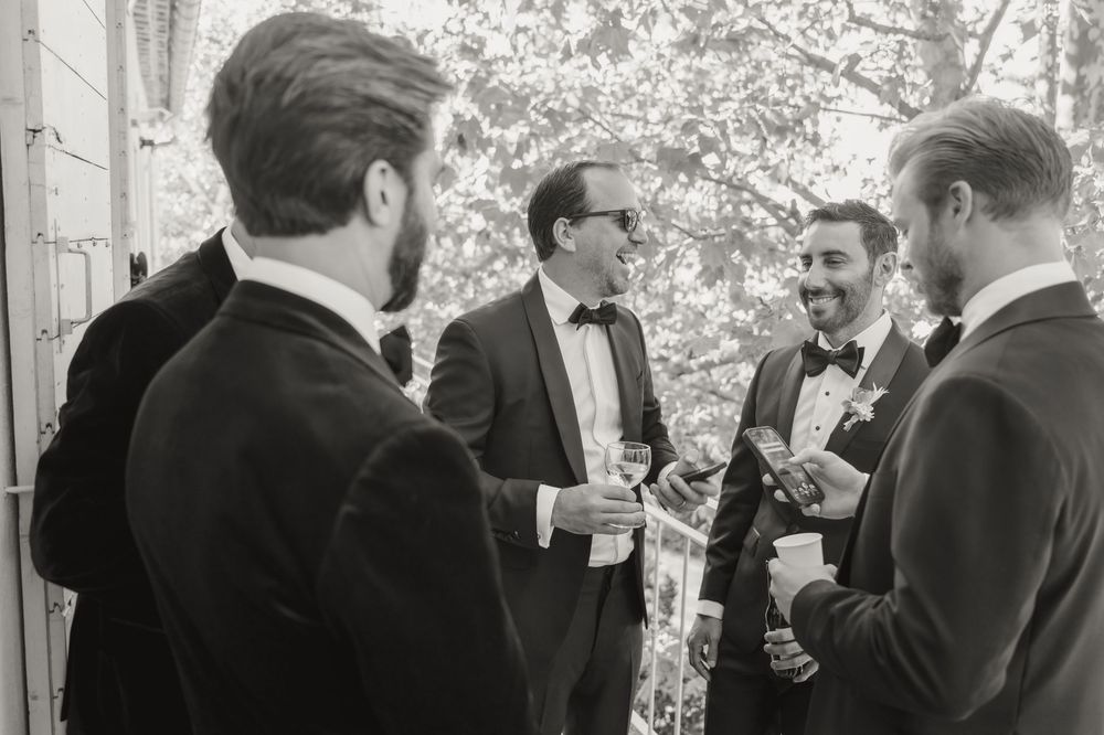 Black and white photo of four men in tuxedos socializing at a formal wedding event.