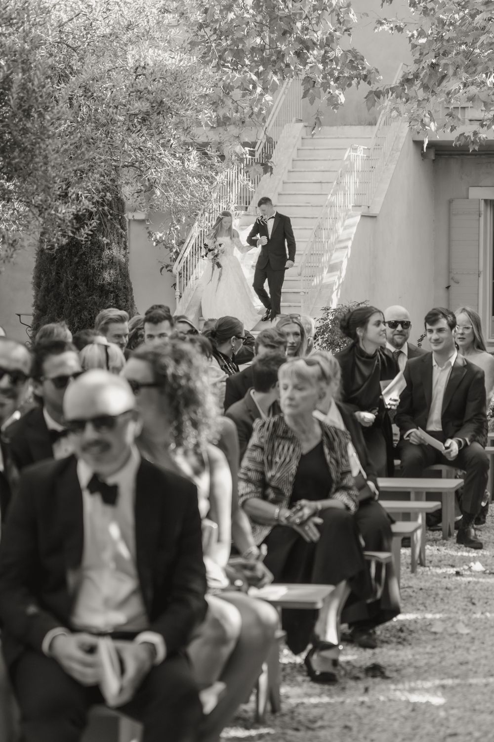 Black and white photo of wedding guests seated outdoors watching bride and groom descend staircase.