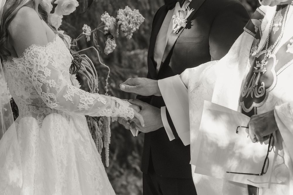 Black and white wedding ceremony photo of bride and groom exchanging rings with officiant outdoors.