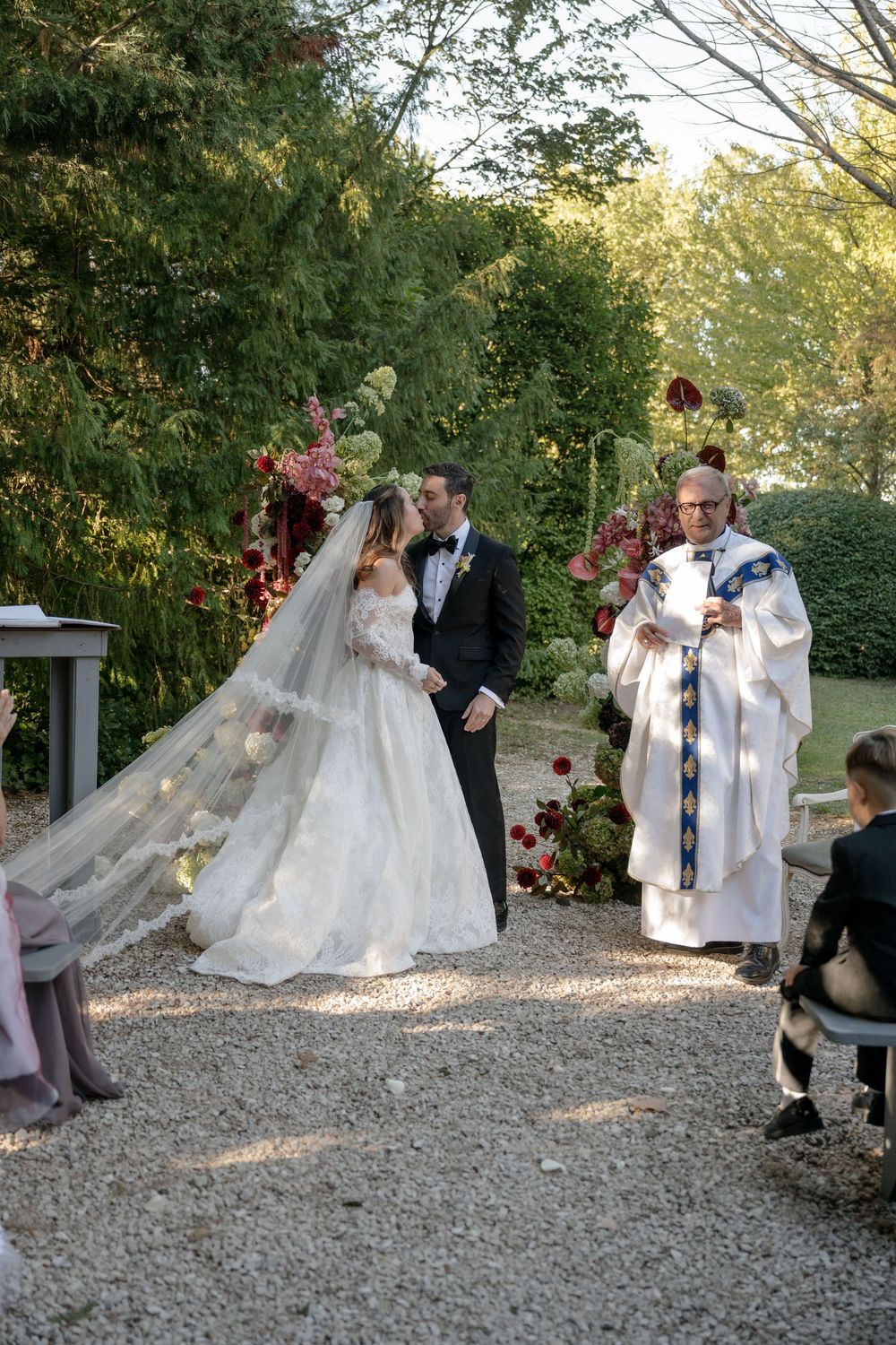 Bride and groom exchange vows outdoors with officiant during elegant garden wedding ceremony surrounded by lush greenery.