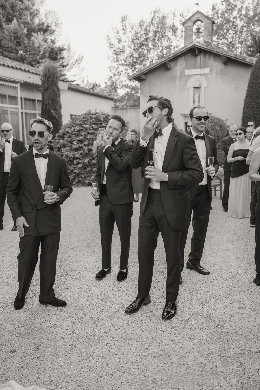 Groomsmen in black tuxedos and sunglasses celebrate outdoors at a wedding venue.