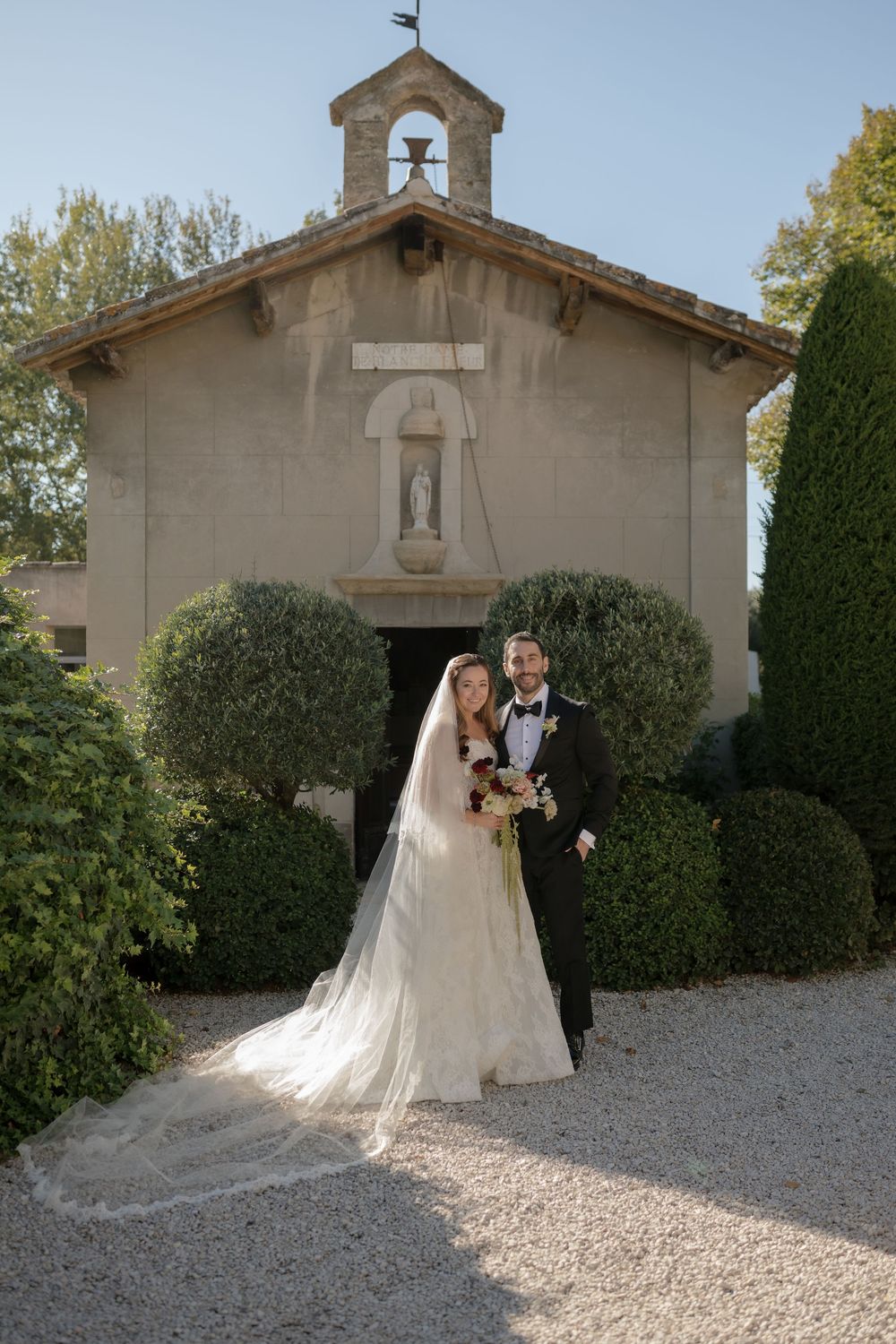 Bride and groom posing in front of a charming stone chapel with manicured hedges.
