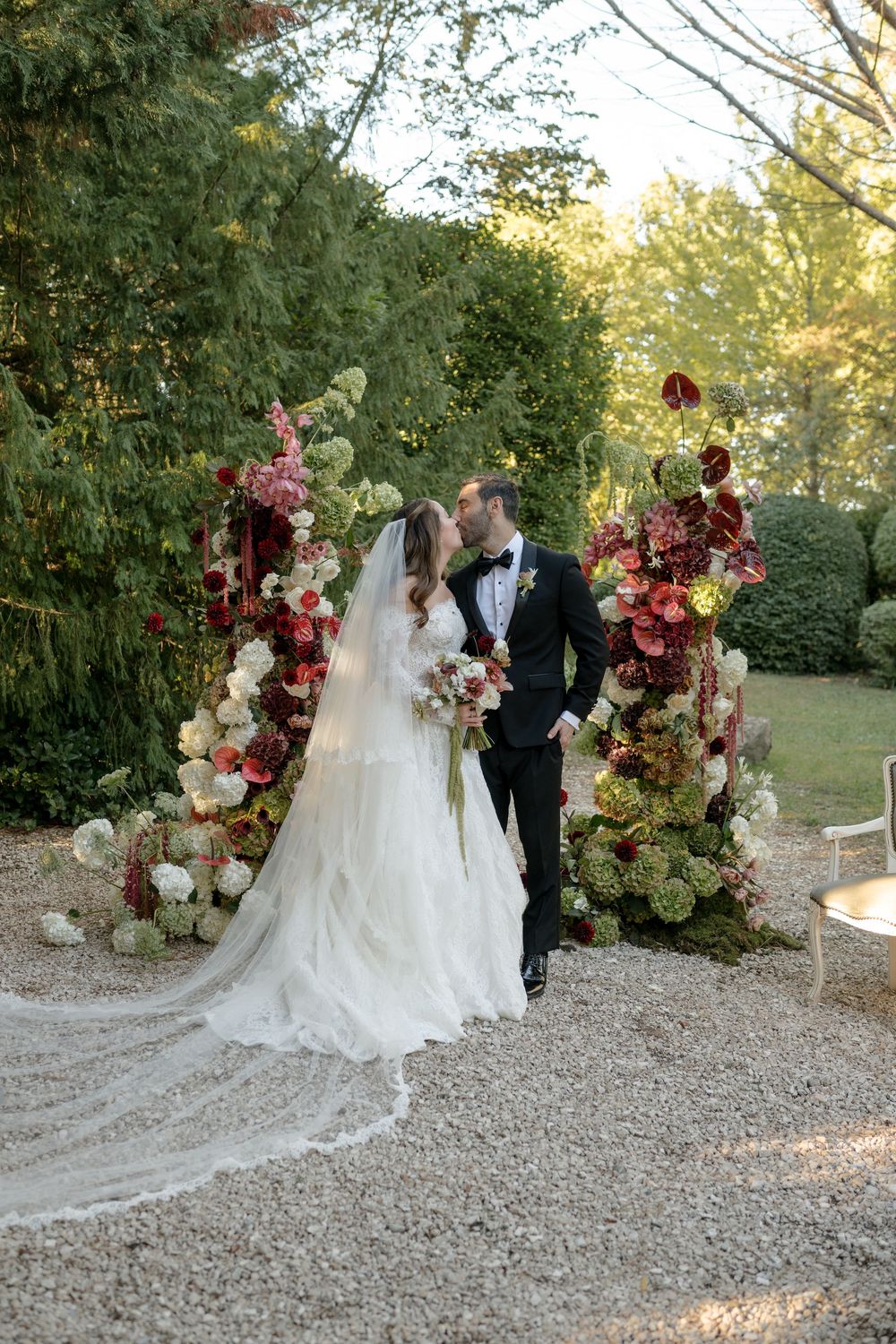 Bride and groom share a kiss beneath an elegant floral arch of red and white blooms.